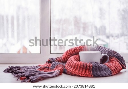 A cup of hot tea warmed in the knitted scarf is on the windowsill against the winter window Royalty-Free Stock Photo #2373814881