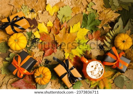 Against the background of autumn leaves there is a ceramic cup with marshmallow dessert, boxes with gifts and cute decorative pumpkins for the Halloween holiday.  Top view, flat lay.