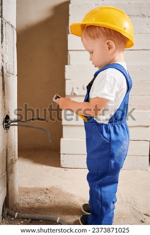 Adorable child construction worker using wire stripper cutter tool while installing electric cables and socket in wall. Kid in safety helmet mounting electrical wiring in apartment under renovation. Royalty-Free Stock Photo #2373871025