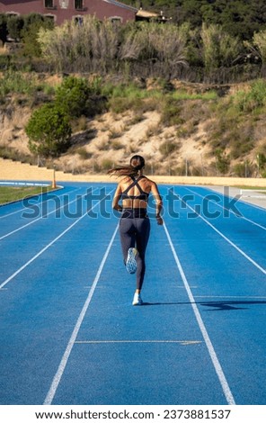 Beautiful tanned slim young runner girl, dressed in tight sportswear, running and sprinting on her back with her tail waving in the wind on a blue athletics track. Royalty-Free Stock Photo #2373881537