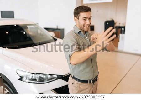 Portrait of cheerful buyer male client doing selfie picture on smartphone at dealership after bought new car. Smiling young man choosing new vehicle in showroom and making photo using mobile phone.
