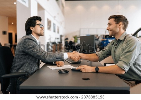 Side view of happy smiling client male purchasing automobile in dealership, signing paper, shaking hands with car dealer in suit enjoying successful agreement. Concept of buying new auto at showroom. Royalty-Free Stock Photo #2373885153