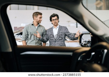 View from interior of luxury vehicle to professional car dealer in business suit showing new car to cheerful male client explaining characteristics. Bearded salesman helping with choice of auto to guy Royalty-Free Stock Photo #2373885155