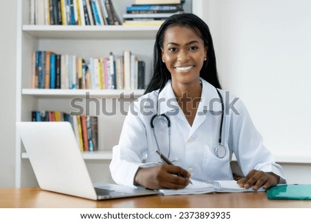 Laughing young latin american female doctor writing notes at desk at office of hospital