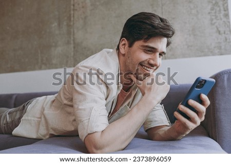 Young side view smiling happy man in casual clothes beige shirt pink t-shirt prop up chin use mobile cell phone chat online lying on grey sofa rest indoors at home on weekends. People leisure concept. Royalty-Free Stock Photo #2373896059