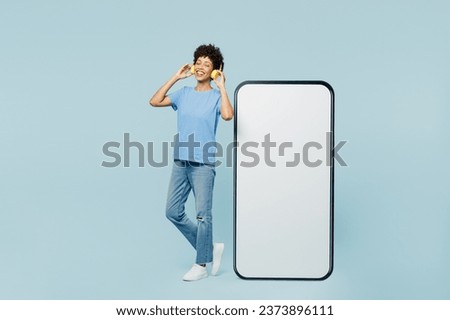 Full body young woman wears t-shirt casual clothes big huge blank screen mobile cell phone smartphone with workspace area listen to music in headphones isolated on plain pastel blue cyan background