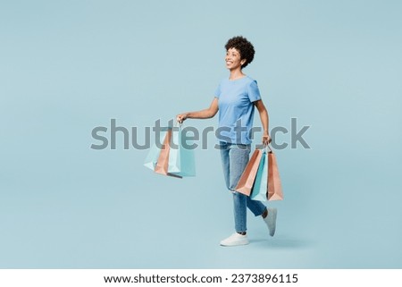 Young woman of African American ethnicity wearing casual clothes hold in hand paper package bags after shopping isolated on plain light pastel blue cyan background. Black Friday sale buy day concept