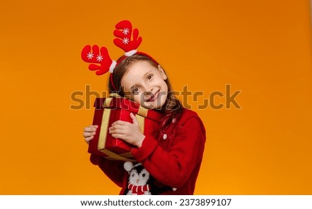 Happy with the gift, a girl in a red Christmas sweater and deer horns on her head holds a gift box in her hands. A girl with wide-open eyes and a sweet smile.