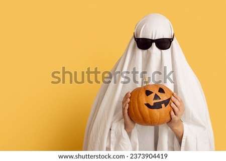 Woman in Halloween costume of ghost and sunglasses holding pumpkin on color background