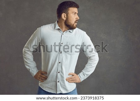 Funny surprised and confused Caucasian man looking around standing on a gray background. Bearded millennial man in a denim shirt holds his hands to his sides and looks away. Concept of confusion. Royalty-Free Stock Photo #2373918375
