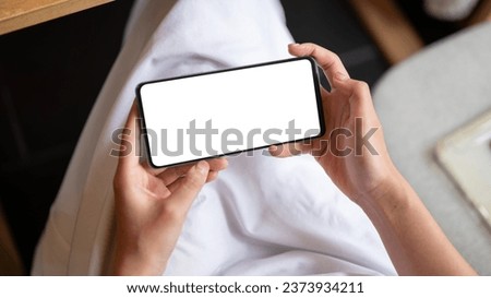 Close-up image of a white-screen smartphone mockup in a horizontal position in a woman's hands. A woman using her smartphone while relaxing in a cafe. watching videos, playing mobile games