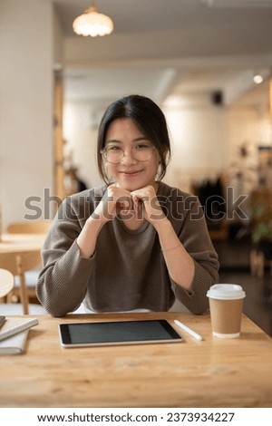 A portrait of a beautiful young Asian woman in casual clothes smiling at the camera and sitting at a table in a beautiful cafe.