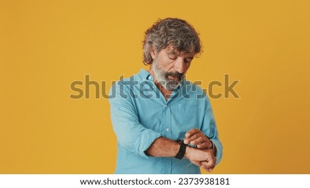 Elderly white-haired bearded man wears a blue shirt, feels bored, looks at his wristwatch isolated on orange background in studio