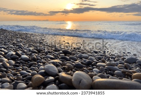 Sunset at sea, view from shoreline of beach with pebbles. Sea beach shore with waves in ocean. Pebble stone beach on sunrise. Shore landscape on Spain resort. Ocean shoreline scenic. Waves in sea.