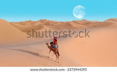 A woman in a red turban riding a camel across the thin sand dunes of the in Western Sahara Desert, Morocco, Africa Royalty-Free Stock Photo #2373944295