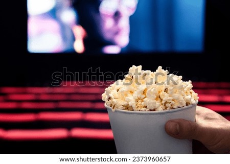 Movies and popcorn. Man holding pop corn box at cinema. Action, thriller or scifi entertainment on screen. Red seats in dark theater. Salty snack in bucket. Spectator pov. Film premiere.