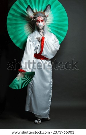 Cheerful woman in animal mask and white kimono standing with green fan on black background, full length portrait. Halloween, Carnival and Cosplay concept