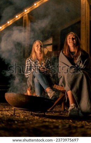 Girlfriends in blankets sit in the evening by the fire near a country wooden house in the forest