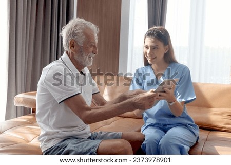 Female nurse takes care of senior patient and teaches him to use digital tablet at living room house, elderly mature grandfather learns to online internet and high-tech, nursing medical health care.