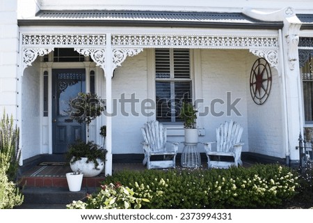 OLD VICTORIAN STYLE TOWN HOUSE FRONT WITH A PORCH VERANDAH A white painted brick house with cast iron decorative trims, a paneled front door patio entrance with timber seats and tiled deck and plants Royalty-Free Stock Photo #2373994331
