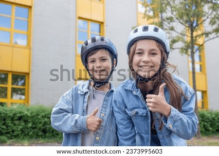 Two children, boy and girl in helmets, stand in a park on the school grounds, background of the institution, children smile, look at the camera, show a thumbs up. Play during recess between lessons.