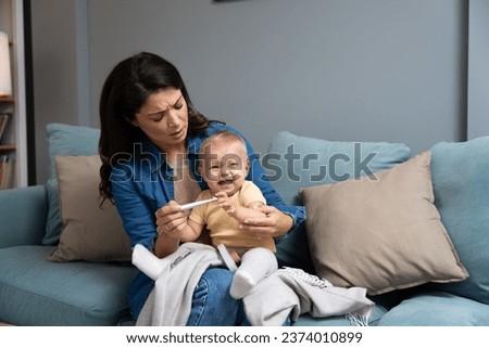 Mother measuring temperature of her sick crying baby sitting on sofa in living room, little infant child catching cold, having influenza or grippe. Young mom worry for her kid cry Royalty-Free Stock Photo #2374010899