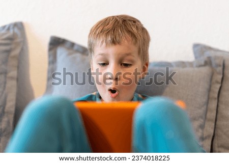 Portrait of cute adorable blond little kid boy enjoy sitting on home sofa surf web play game or watching TV show movies using tablet. Children smart technology usage concept. Child gadget addiction