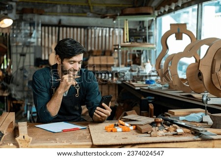 A luthier man crafts an acoustic guitar in his workshop, surrounded by tools and materials, while writing on white paper and texting a customer on his smartphone.