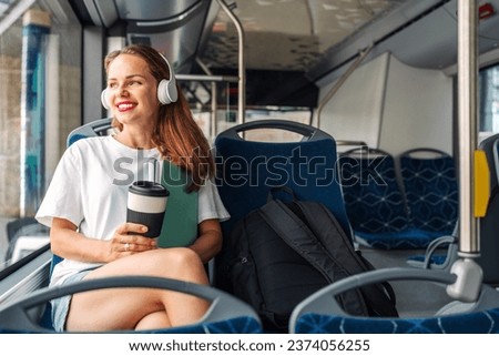 Attractive young adult woman listening music while traveling by city shuttle bus, holding travel mug in hands. Royalty-Free Stock Photo #2374056255