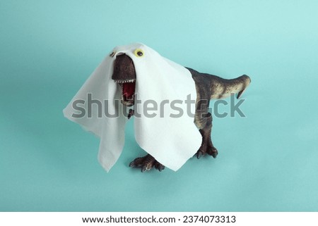 a plastic t-rex dinosaur figurine disguised as a ghost with a sheet and false eyes on a turquoise background. Minimalist, trendy still life photography.