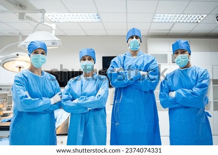 Selective focus of a professional surgical team, with one male and two female Caucasians, and Asian woman, in blue surgical uniforms, caps, and face masks, standing with arms folded looking at camera. Royalty-Free Stock Photo #2374074331