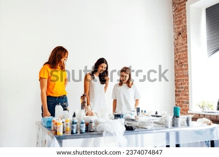 Cute teenager in white T-shirt and two women in casual clothes are checking out acrylic-painted work. Young artist communicates with teachers in art workshop. Paints, brushes and cups on table.