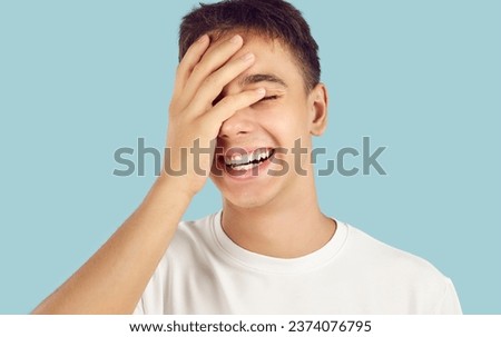 Portrait of a young cheerful laughing smiling boy student in casual clothes covering his eyes with hand standing isolated on a studio blue background. People emotions and lifestyle concept.