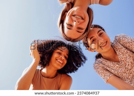 Funny multiracial smiling teenager gilrs standing and looking down at camera. Group of happy cheerful female friends in casual clothes having fun together outdoors. Low angle view. Copy space. Royalty-Free Stock Photo #2374084937
