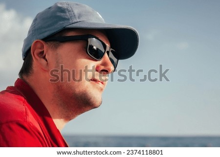 portrait of young man in sunglasses, red t-shirt and blue cap on sea shore background. Travel vacation holiday