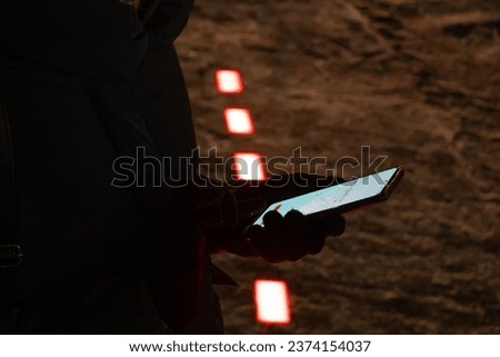 Women's gloved hands hold a mobile phone waiting for a green signal at a ground traffic light in the winter evening. New modern traffic lights built on the sidewalk for pedestrians