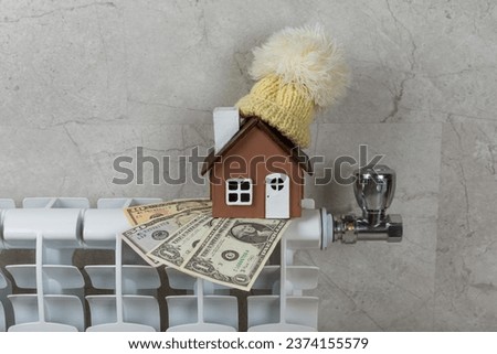 Home heating radiator. Model of a house wrapped in a scarf and hat on a radiator indoors against a wall. Efficiency of winter heating. Concept of heating season. space for text.Copy space.