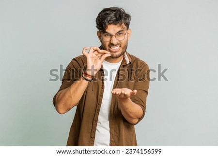 Need some more, please give me. Indian man showing a little bit gesture with sceptic smile showing minimum sign, measuring small size, begging help. Arabian guy isolated on gray studio background Royalty-Free Stock Photo #2374156599