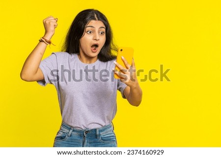 Happy excited Indian woman girl use smartphone typing browsing screaming say wow yes found out great win good news lottery goal achievement celebrating success, winning play game on yellow background Royalty-Free Stock Photo #2374160929