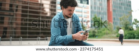 smiling young bearded man in denim shirt using mobile phone. Man looking at his smartphone social networks on modern city background