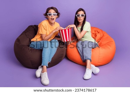 Full body photo of girls together watching barbie new film cinema eat yummy popcorn sit vip bean bags isolated on purple color background