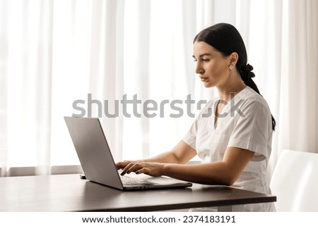 Close-up of a female family doctor working on a laptop computer in a clinic. A doctor in a white lab coat reviews a medical history at a table in a hospital office.