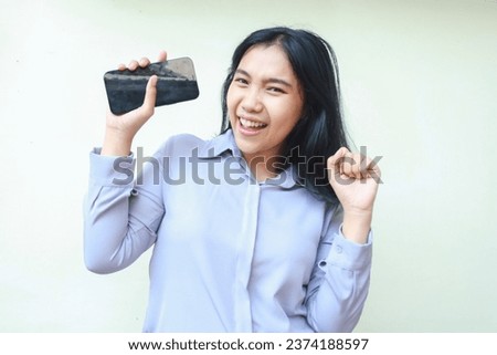excited asian young business woman holding mobile phone and raising fist pump say yes gesture winning achievement, celebrate victory with dancing, wearing formal shirt standing over white background