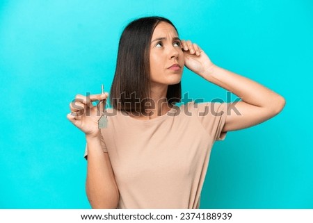 Young woman holding home keys isolated on blue background having doubts and with confuse face expression Royalty-Free Stock Photo #2374188939