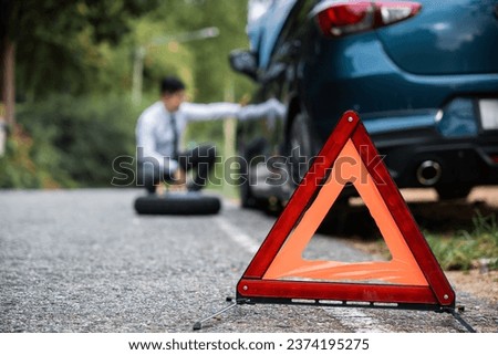 Close up of Red triangle sign behind the broken car. Side view of Businessman sitting on road sign wheel changing. Warning triangle in front of broken-down car on the road.