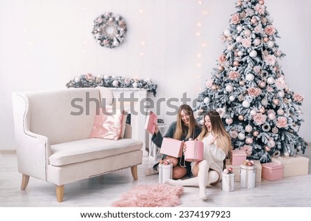 Smiling young women opening a gift box near beautiful Christmas tree at home. Presents for Christmas and New Year. white room with fireplace and sofa.