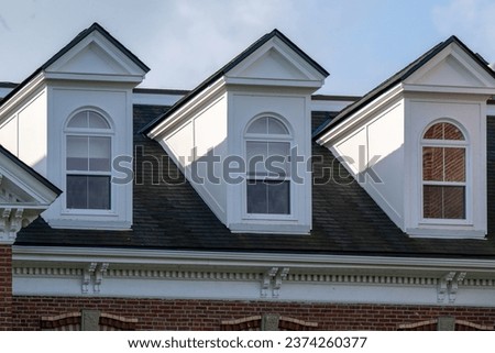 Three peaked dormer-style windows on a hip roof covered with black shingles. The vintage white wooden frames have arch-shaped glass and closed double-hung panes with white curtains hanging inside.   Royalty-Free Stock Photo #2374260377