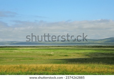 Endless flat steppe with low grass under a cloudy sky and a wide lake in the background at the foot of a range of hills. Khakassia, Siberia, Russia.