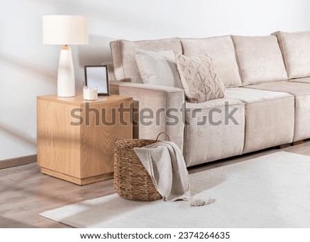 Living Space with Beige Upholstered Sofa with Textured Pillows, Wooden Side Table Hosting Ceramic Lamp, Picture Frame, Woven Basket with Throw Blanket, Set on Light Wooden Flooring and White Rug.