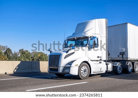 White classic industrial local carrier day cab big rig semi truck tractor with roof spoiler transporting commercial cargo in dry van semi trailer running on the wide highway road at sunny day Royalty-Free Stock Photo #2374265811